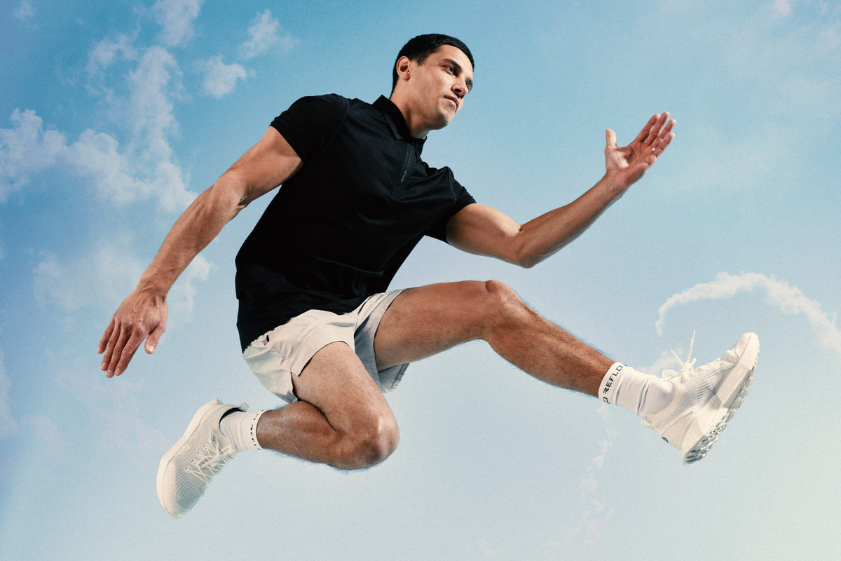 A man in a black polo shirt, white shorts, and white sneakers jumps through the air against a bright blue sky with light clouds.