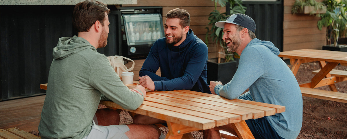 Three men sitting at a wooden picnic table outdoors, engaged in conversation wearing Reflo clothing.